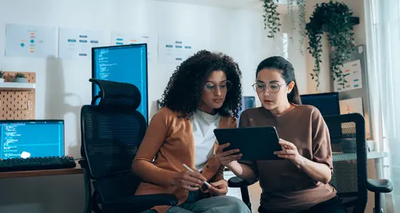 Women in tech collaborate on UI review with tablet and coding monitors in background