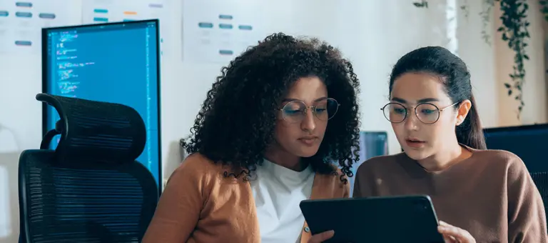 Women in tech collaborate on UI review with tablet and coding monitors in background
