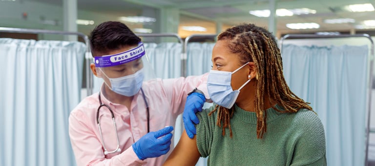 A woman wearing a facemask receiving a vaccine from someone in PPE