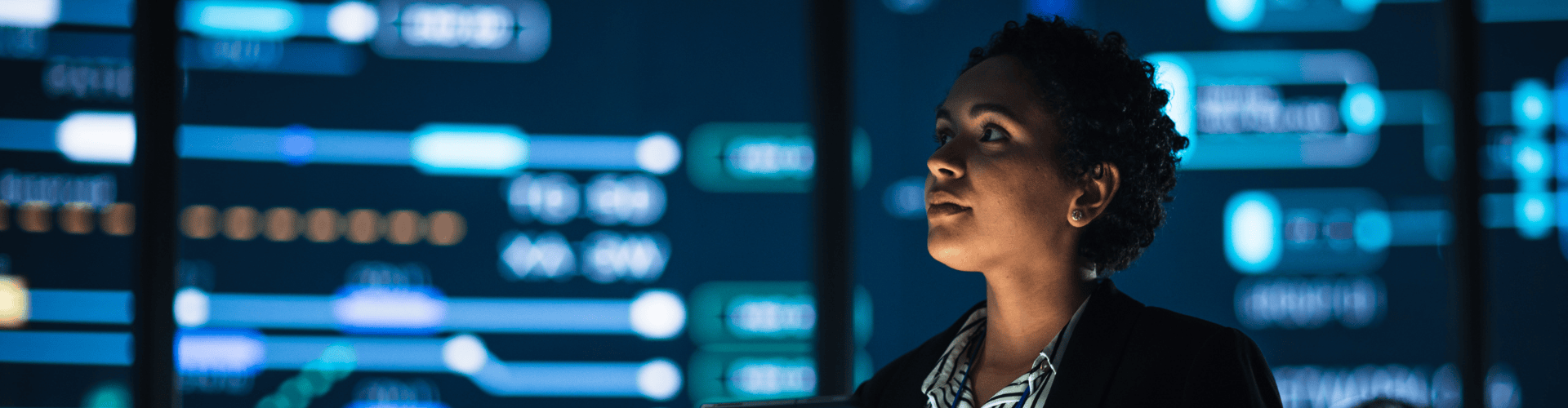 A woman in a blazer holds a tablet, looking up at a wall of glowing data and network diagrams.