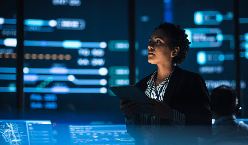 A woman in a blazer holds a tablet, looking up at a wall of glowing data and network diagrams.