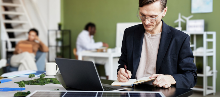Young businessman making notes in notebook while working at table with laptop and solar panel