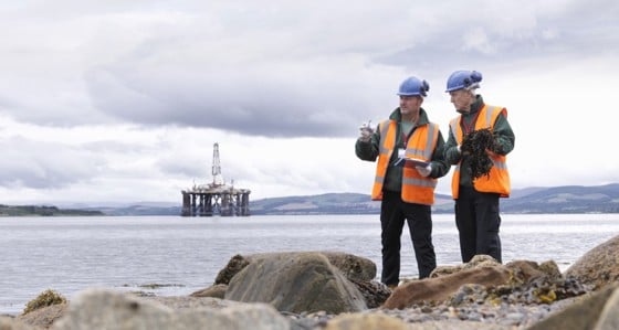 Two engineers standing on the shoreline with an oil rig in the background