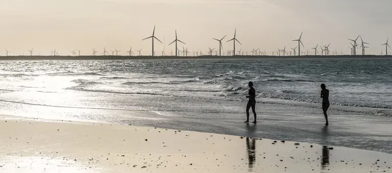 Two people walking on a beach with a windfarm in the distance