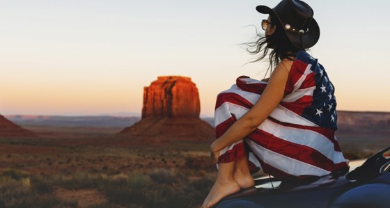A woman sitting in the desert draped in the Stars and Stripes flag
