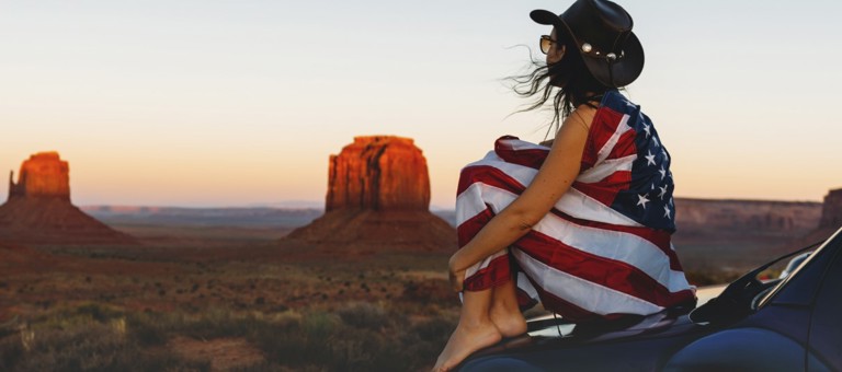 A woman sitting in the desert draped in the Stars and Stripes flag
