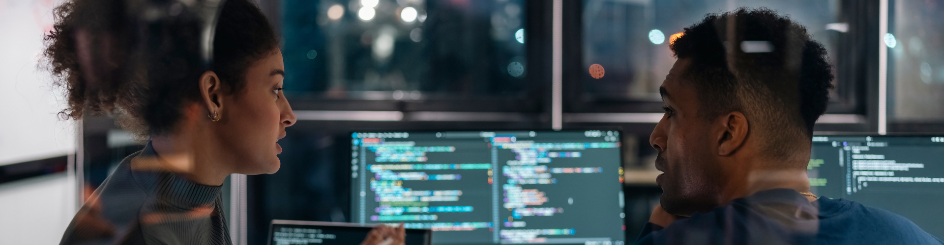 A woman and man work together on laptops at night in an office with code on the screens.
