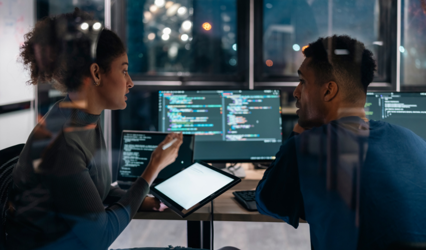 A woman and man work together on laptops at night in an office with code on the screens.