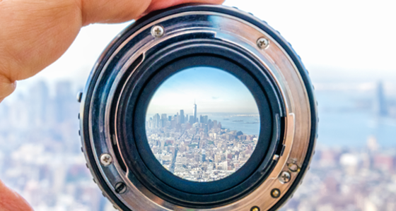 A hand holding a camera lens with a view the Lower Manhattan cityscape