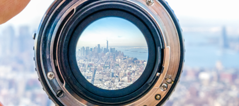 A hand holding a camera lens with a view the Lower Manhattan cityscape