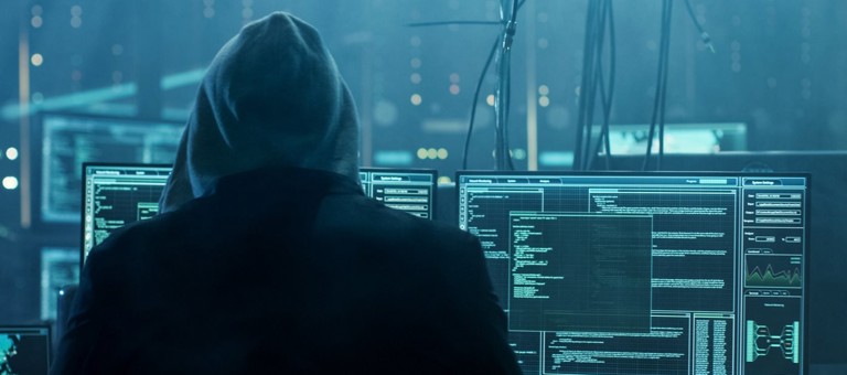 A hacker whose identity is hidden by a hooded top looking at multiple screens