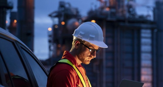 A man in hard hat looking at a tablet in front of industrial buildings at night