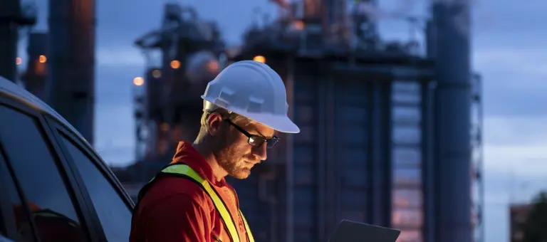 A man in hard hat looking at a tablet in front of industrial buildings at night