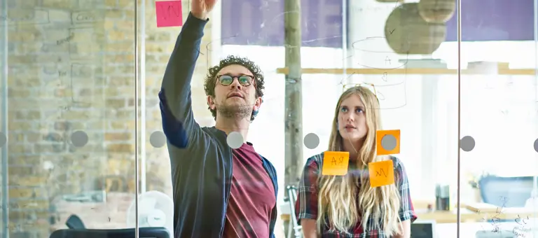A man writing data on a perspex wall with colleague watching