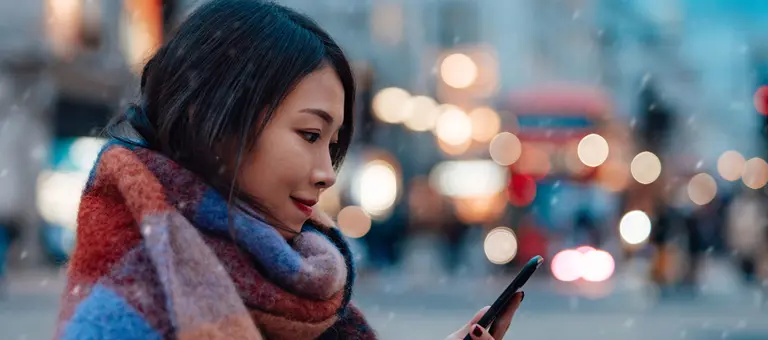 A woman wearing a scarf looking at her phone standing outside while sleet is falling
