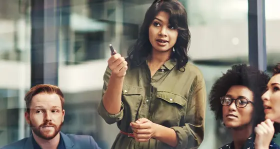 A woman standing up in a group of people pointing with her pen