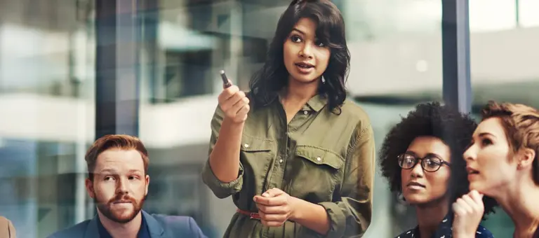 A woman standing up in a group of people pointing with her pen