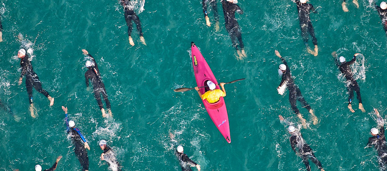 A pink kayak paddling among wet-suited sea swimmers