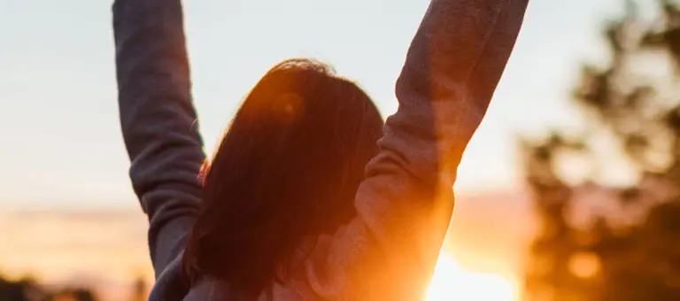 Rear view of young woman stretching arms against sunset in the sky