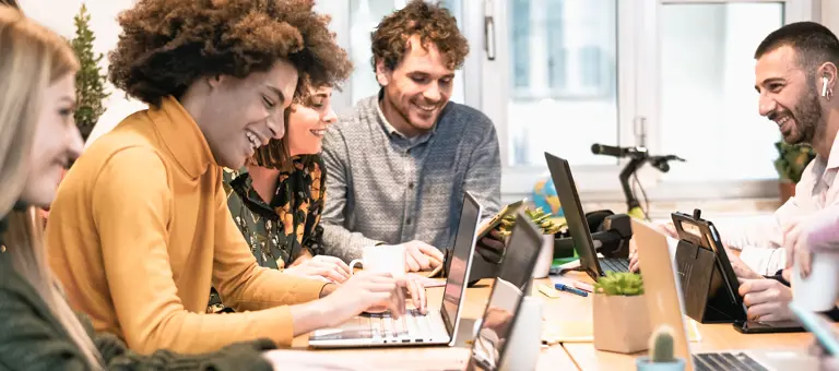A group of laughing and smiling interns sitting around a table with their laptops