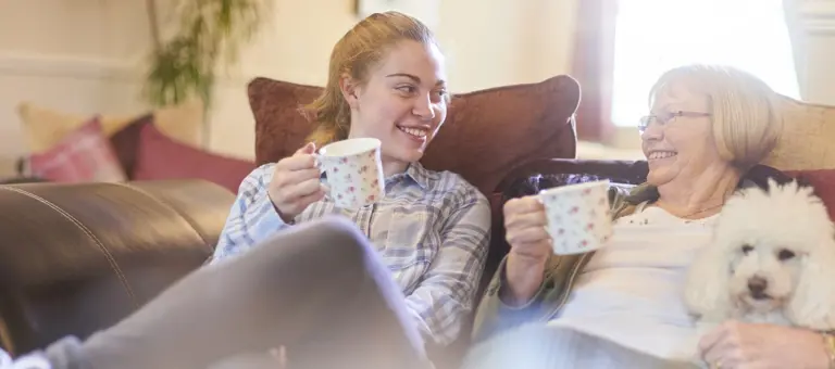Two women drinking cups of tea sitting on the sofa next to their dog