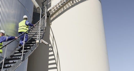 Two men in overalls and hard hats climbing up stairs in a oil storage facility