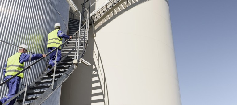 Two men in overalls and hard hats climbing up stairs in a oil storage facility