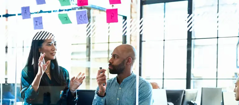 A man and woman discussing post-its on a transparent office wall