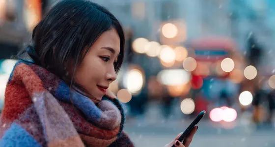 A woman wearing a scarf looking at her phone standing outside while sleet is falling